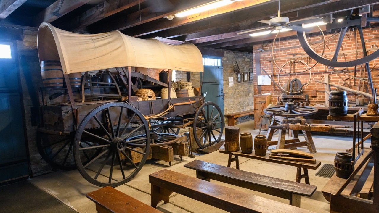 A covered wagon sits inside a room with simple benches in the foreground and many wagon wheels and tools in the background.