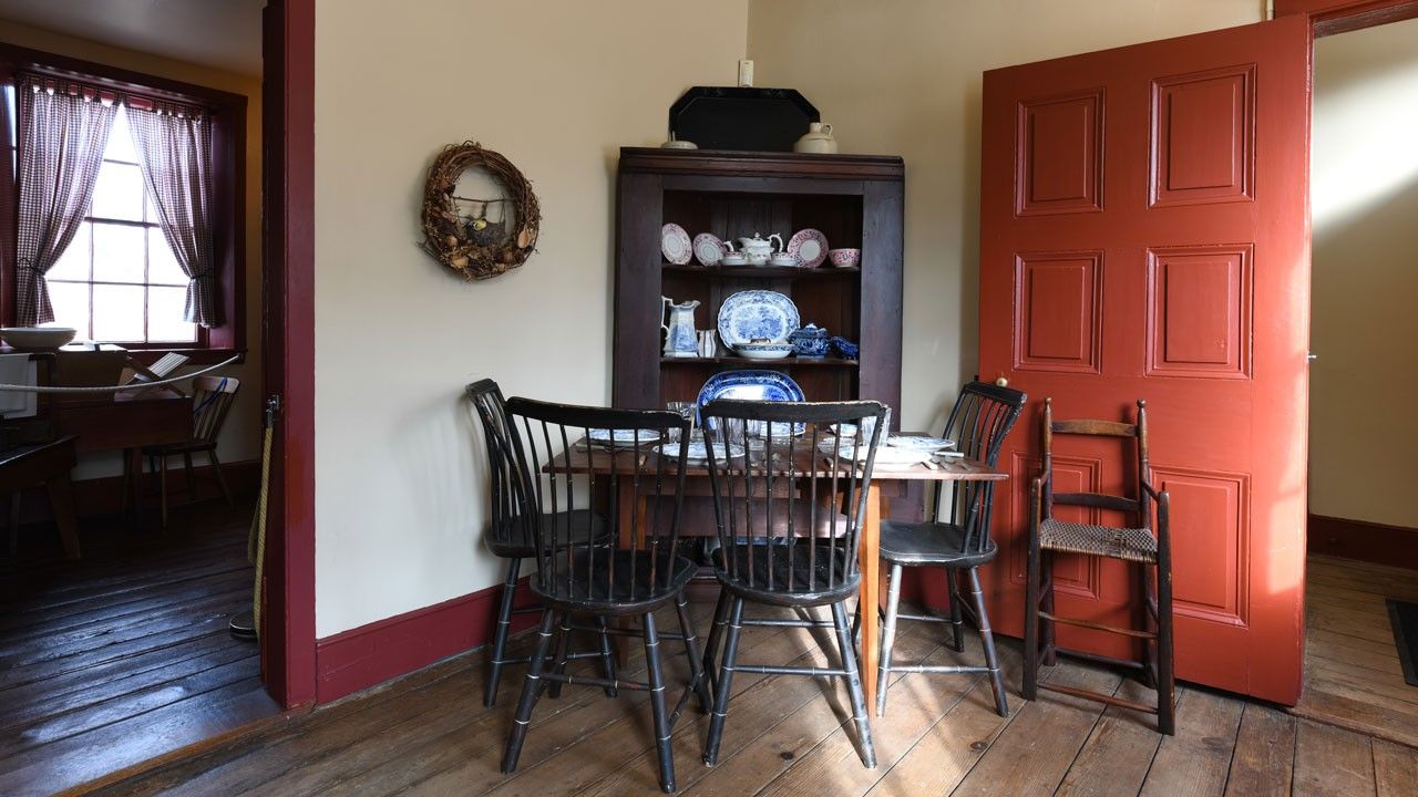 A dining room interior with wood floor and chairs surrounding a table with a corner China cabinet and a red door behind. 