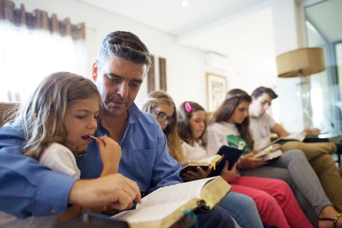A man in a blue shirt sits with five of his children on a long couch to read the scriptures together.