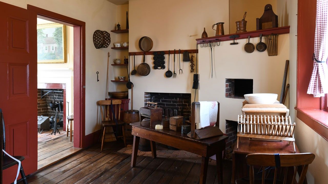 A kitchen interior with wood floor and many cooking utensils and implements hanging on walls, shelves, and tables. 