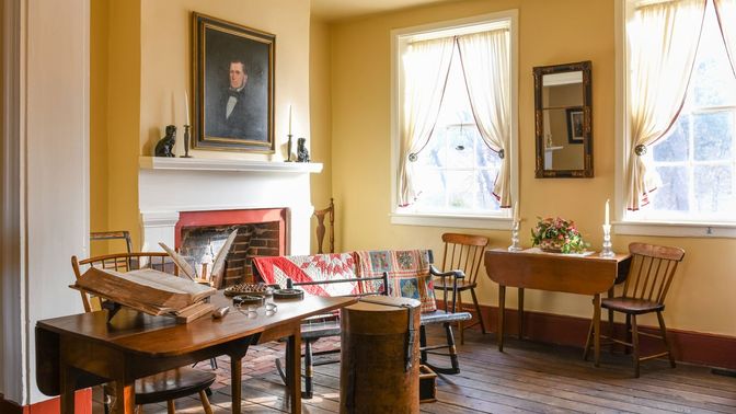 A room with a fireplace, chairs, a large hatbox on the ground, and a open book and quills on a nearby table.