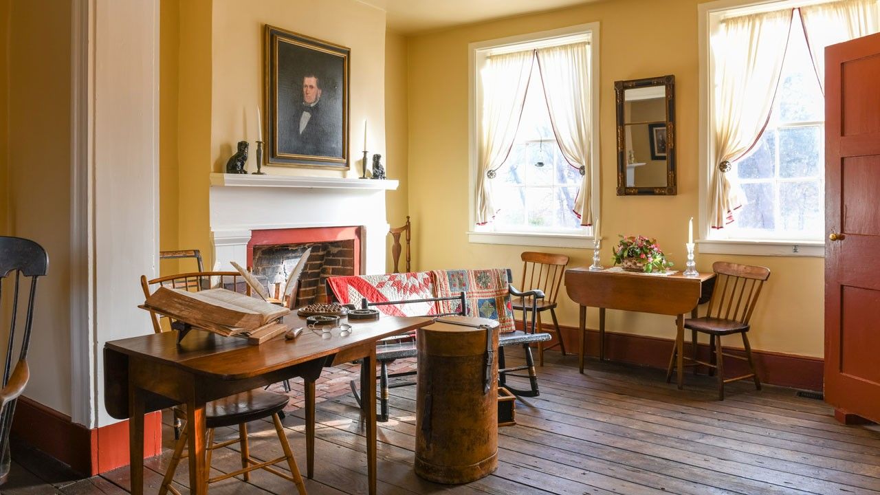 A well-furnished double parlor interior with a portrait painting of a man above a fireplace and bright curtained windows to the right.