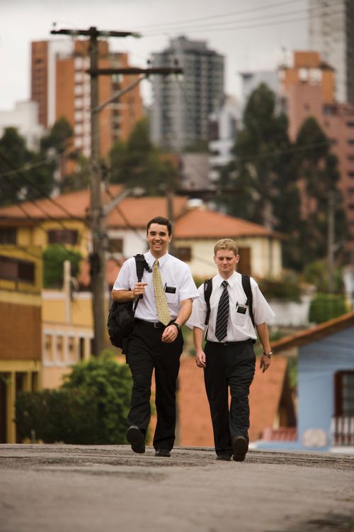Two elder missionaries carrying backpacks and walking up a street in São Paulo, Brazil, with houses, trees, and tall buildings in the background.
