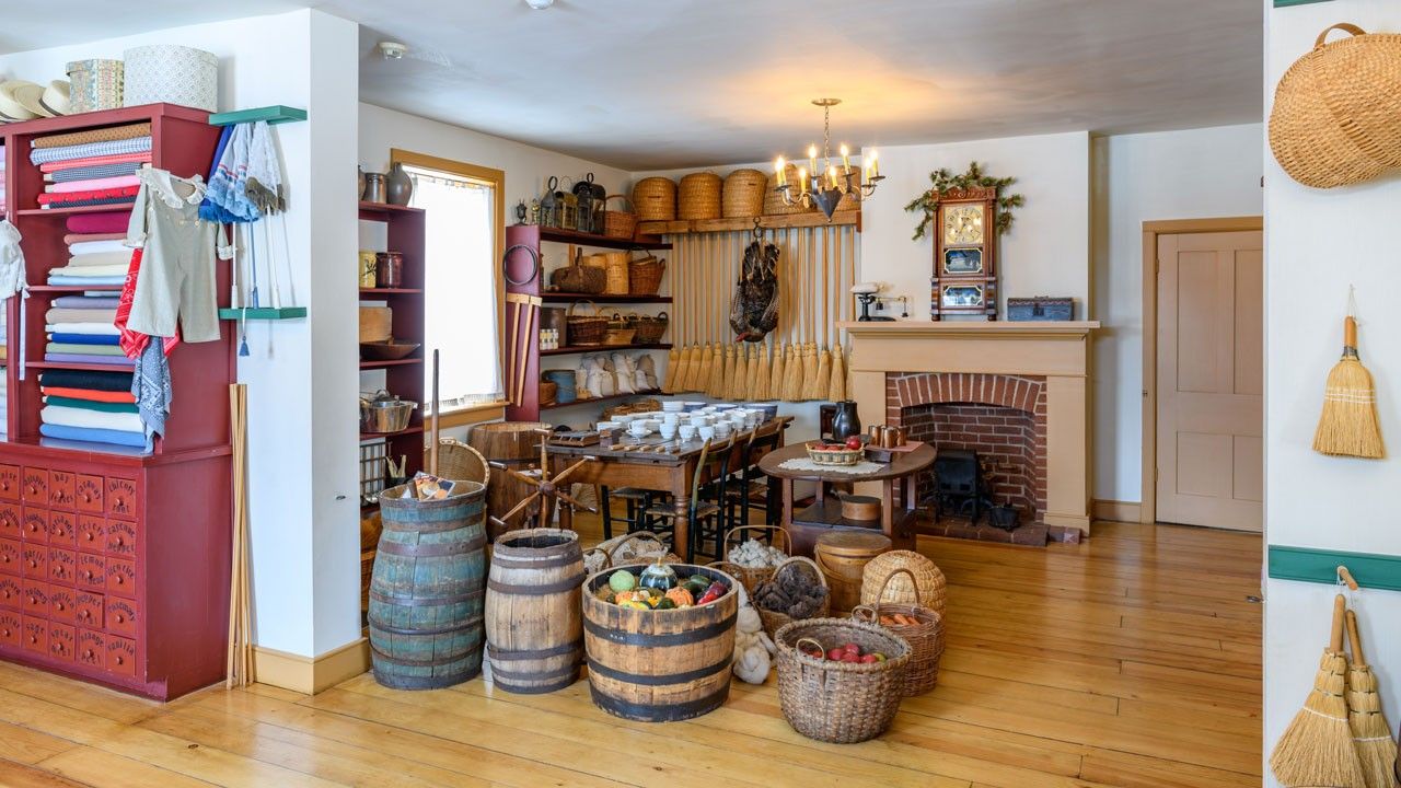 A store with miscellaneous goods, such as brooms and dishware. There are also barrels and baskets filled with wool and food.