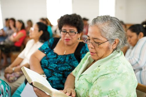 Two women sit next to each other  in a chapel meetinghouse, holding scriptures.