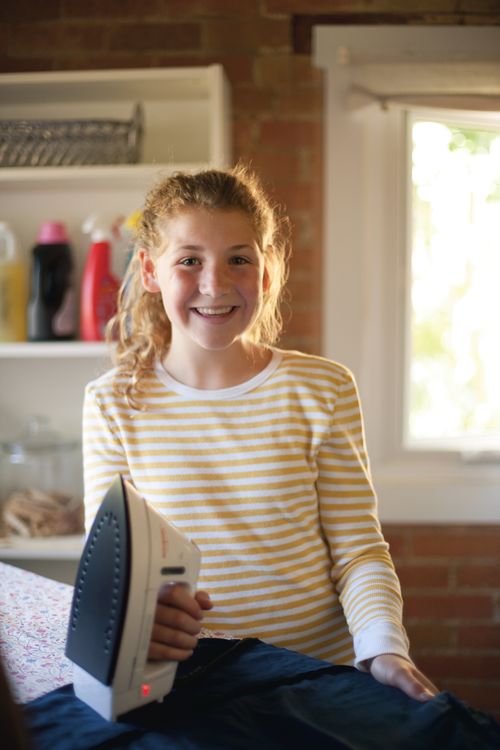 A young woman in a striped shirt smiles and stands at an ironing board while holding an iron.