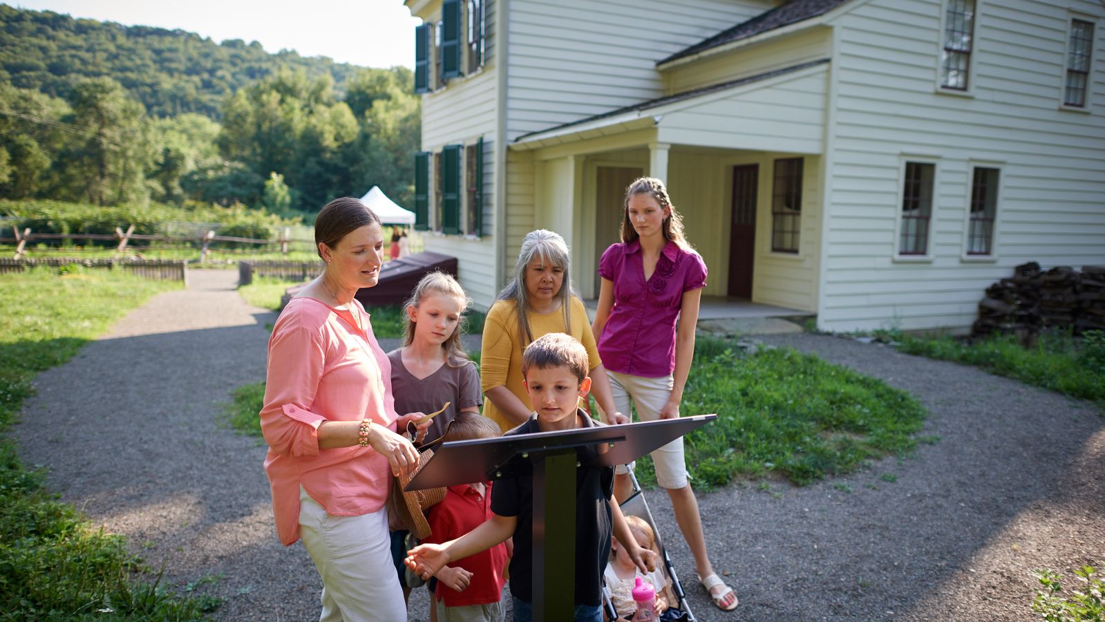 Various people interact with exhibits in Historic Sites found in New York and Pennsylvania. A family stands outside a white home and is looking at a plaque.