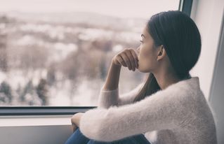 Picture of a woman sitting next to a window. She is looking outside and it looks like she's thinking.