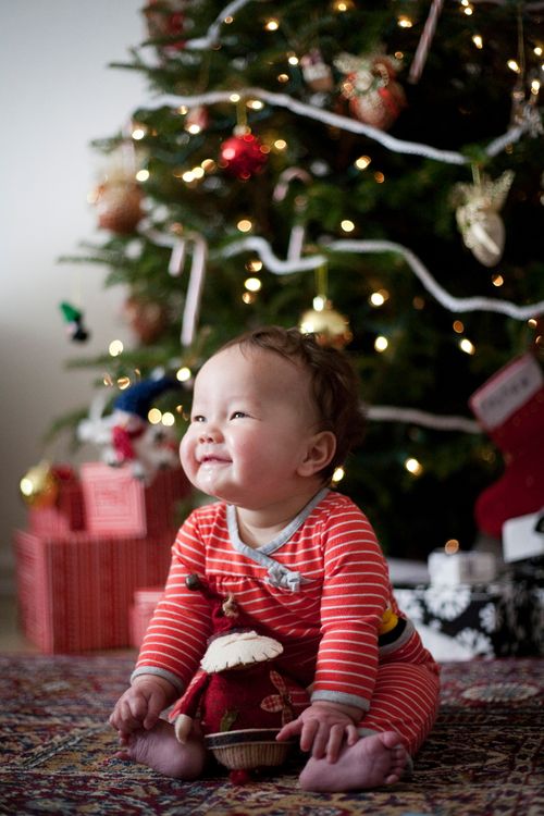 A small baby in red and white striped pajamas smiling and sitting near a decorated Christmas tree in the morning.