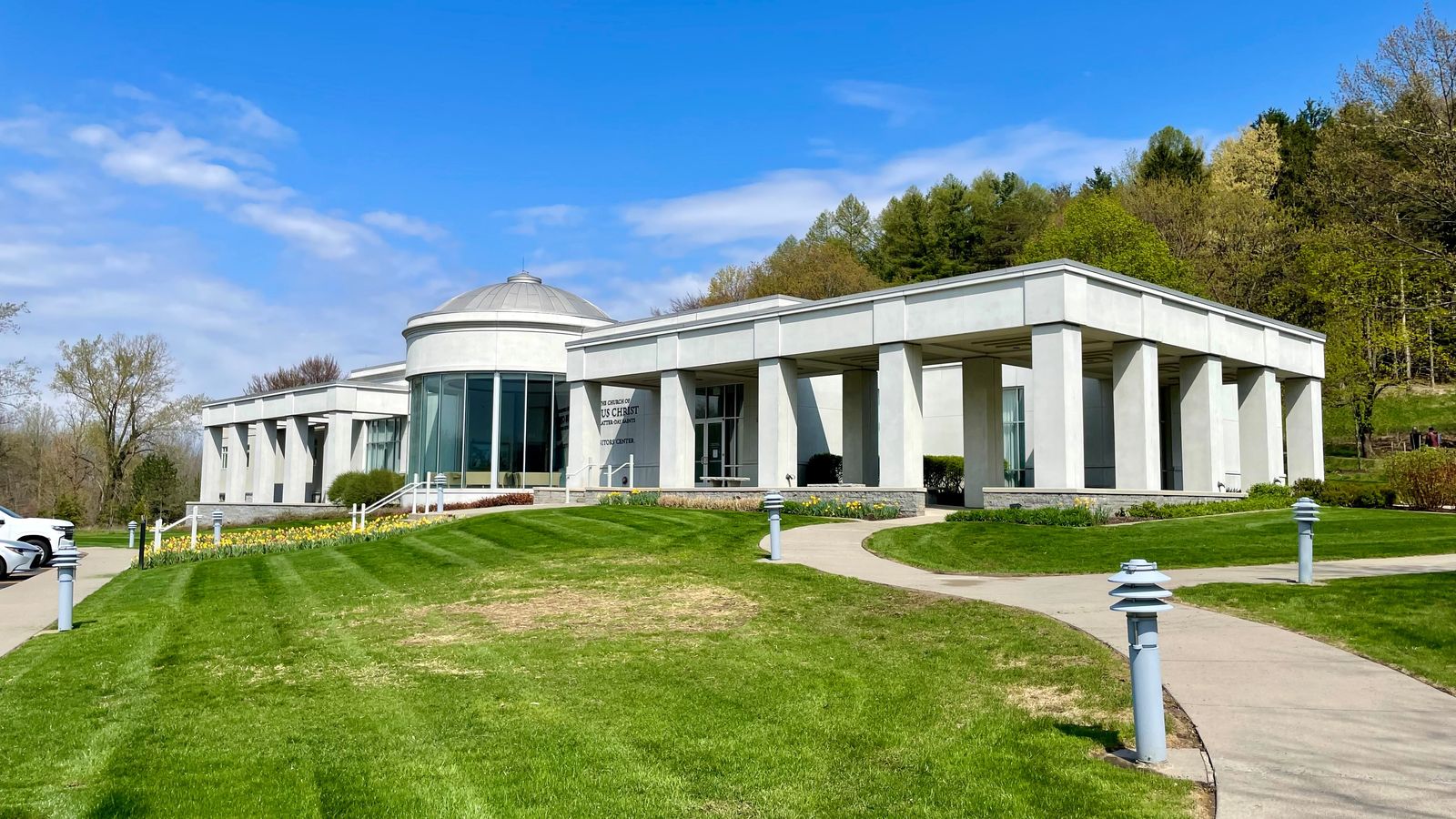 View of a large white building round glass rotunda in the front. A concrete path through a green lawn leads up to it.