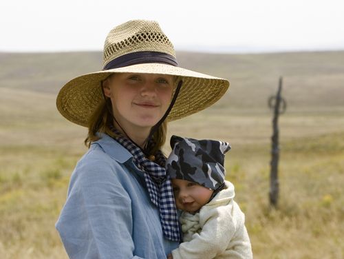 A young woman wearing a wide-brimmed straw hat, a blue button-up shirt, and a scarf stands outside in a grassy field and holds a little baby.