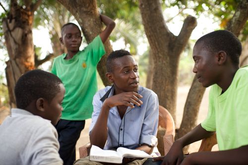 Four young men gather outside by trees and discuss the scriptures together.