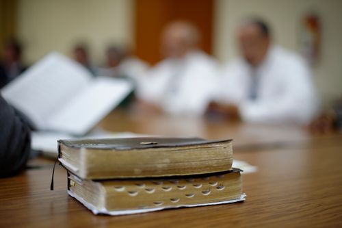 A set of well-used black leather-bound scriptures on a desk, with a classroom setting seen in the background.