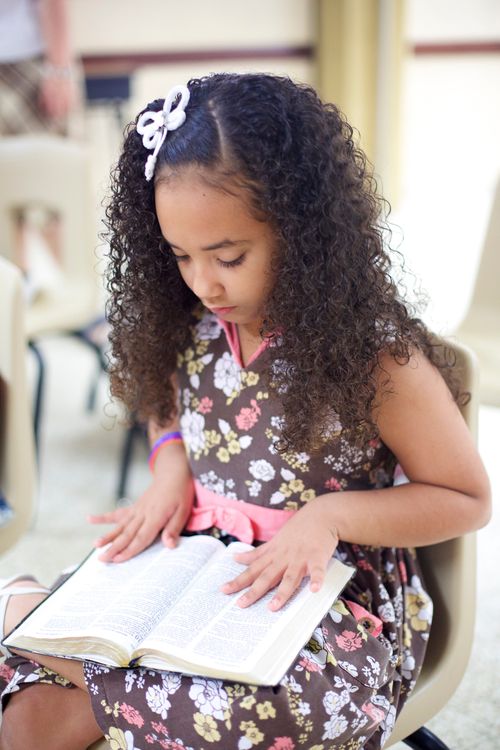 A little girl in a dress sits in a plastic chair and reads the scriptures that she holds on her lap.