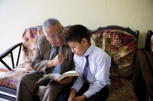 A man and a young boy sit on a couch in Sunday clothing and read the scriptures together.