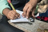 Hand of mother with coins, teaching son who is filling out tithing slip, to pay tithing, in Mumbai, India. (horiz)