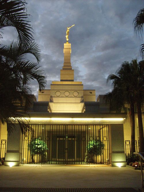 A view of the gated door of the Brisbane Australia Temple lit up at night, with palm trees on the side.