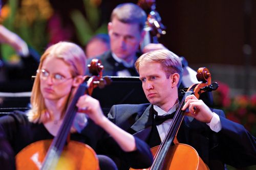 A man in a black tuxedo plays a cello onstage in the Conference Center, with one cellist in front of him and another behind him.