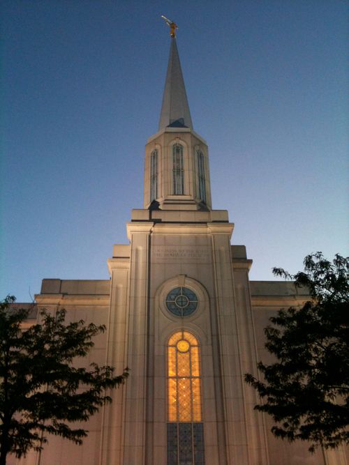 A front view of the St. Louis Missouri Temple in the evening, with trees on either side and the windows lit up from the inside.