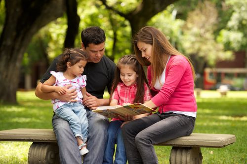A mother and father sit on a bench with their two children and read a book to them.