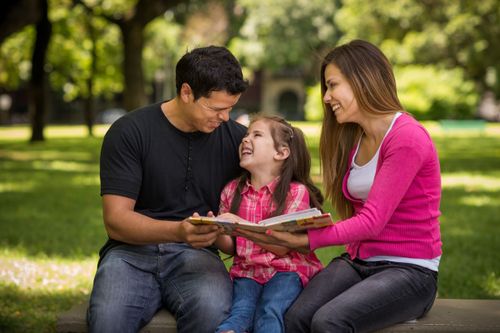 A mother and father sit on a bench in the park, with their daughter sitting between them, and look through a family album.