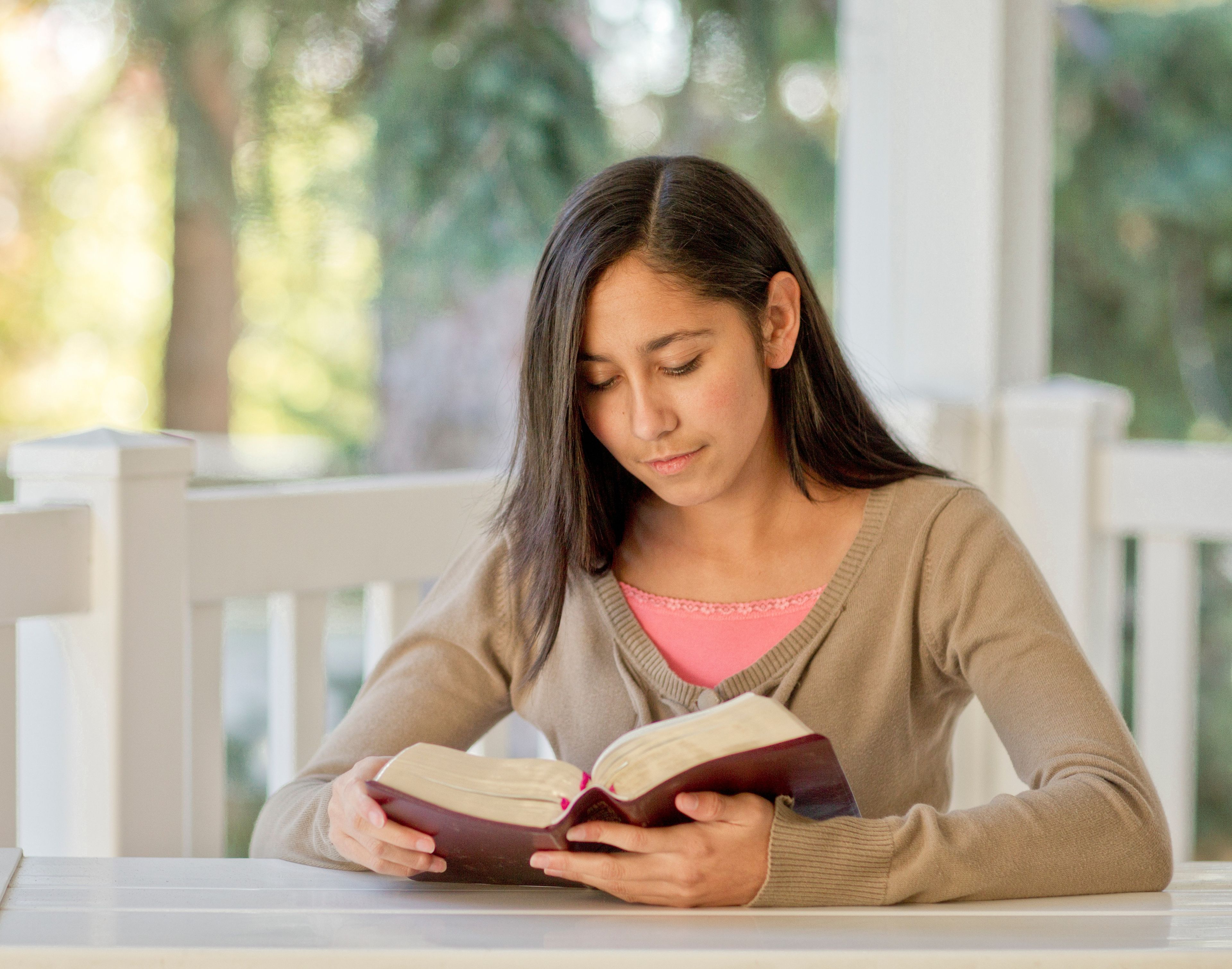 A young woman sits at a table on her porch and reads her scriptures.  