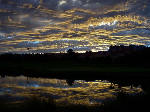Clouds in the sky are reflected in a pond just after the sunset, with large houses and green grass in the background.