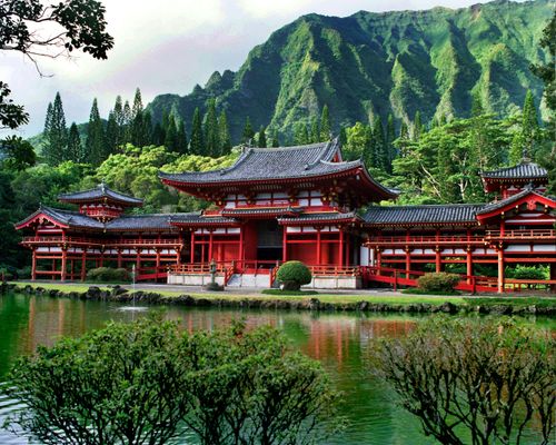 A large, white, black, and red Buddhist temple in Hawaii, seen beyond a pool of water with large green trees and mountains rising in the background.
