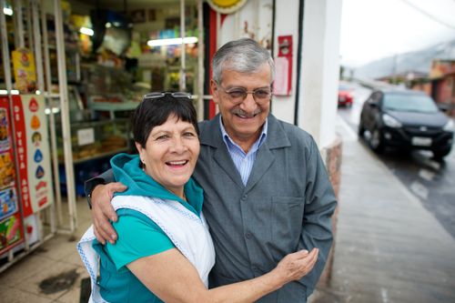 An elderly husband and wife hug each other outside on the sidewalk near a store, with cars seen in the background.