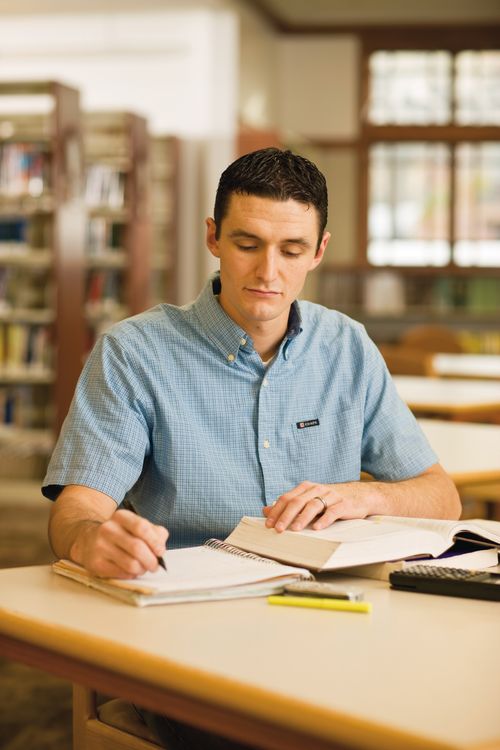 A young man in a blue button-up shirt sits at a table in a library and writes notes in a spiral notebook while reading from a large textbook.