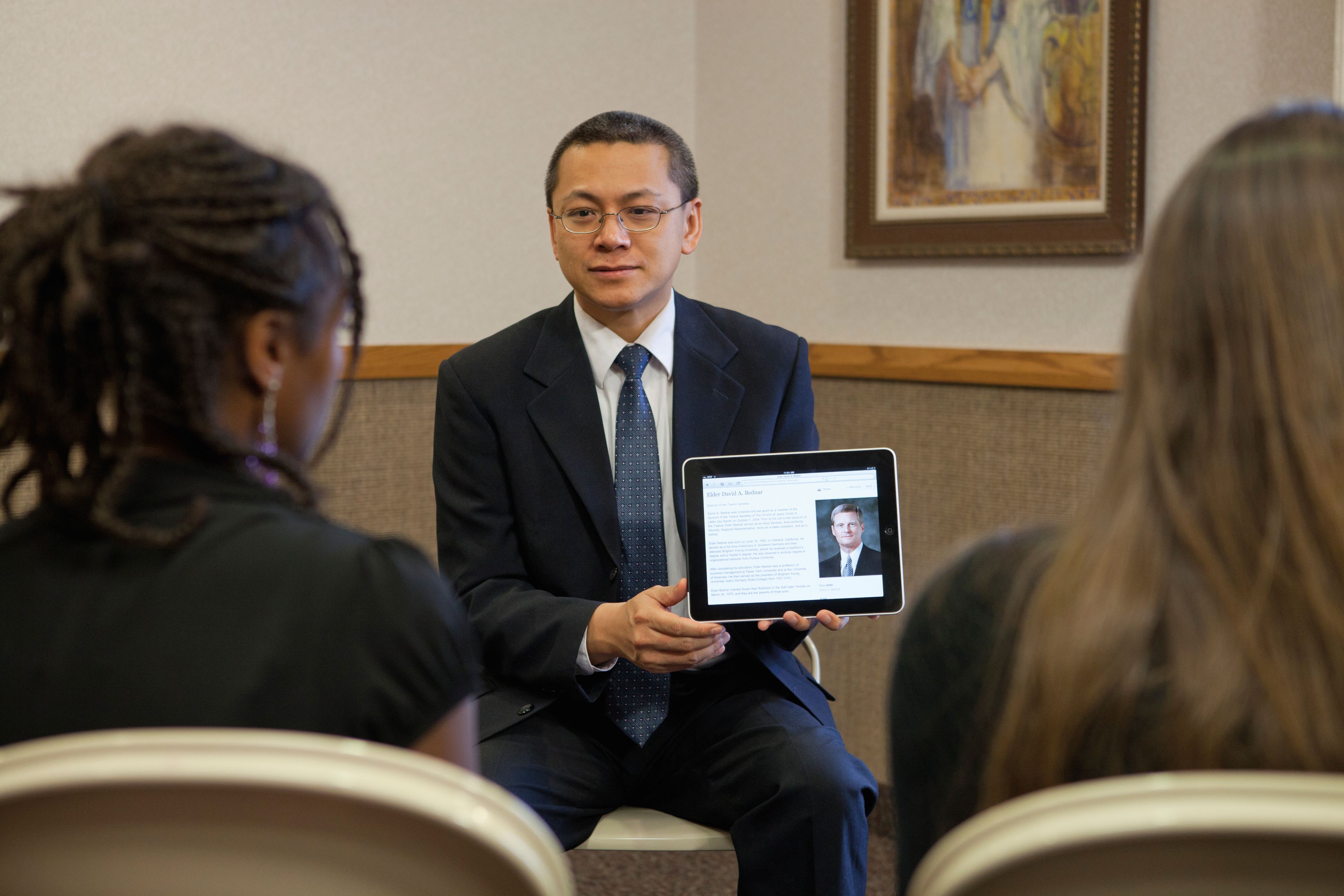 A teacher sits in front of his class and uses a tablet to show his students a talk by Elder Bednar.