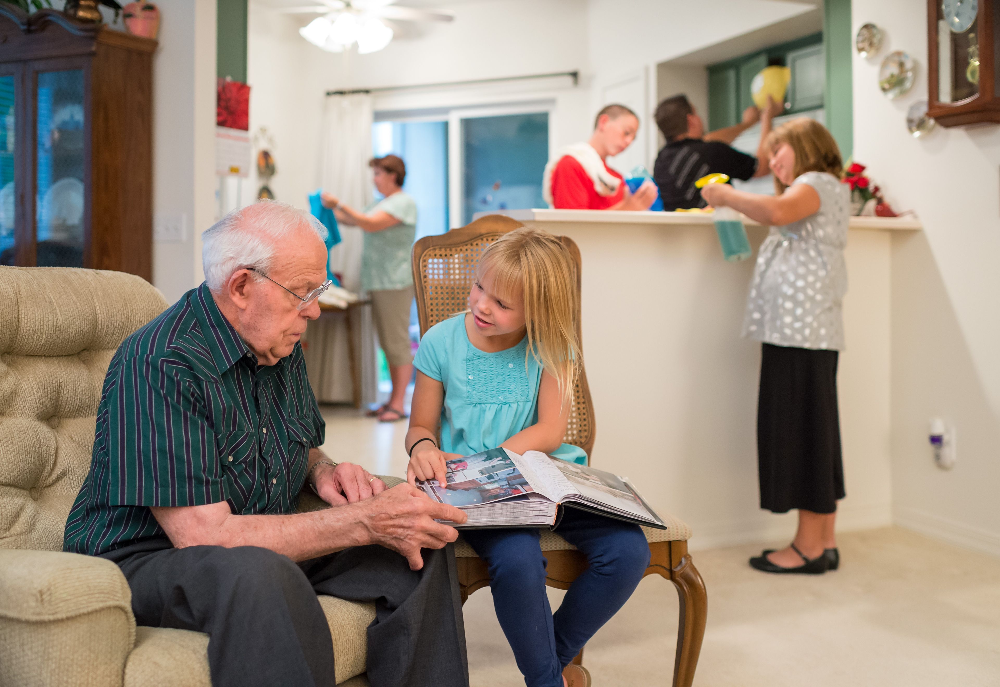 A little girl reads a book to her grandfather while the rest of her family members clean up his house.  