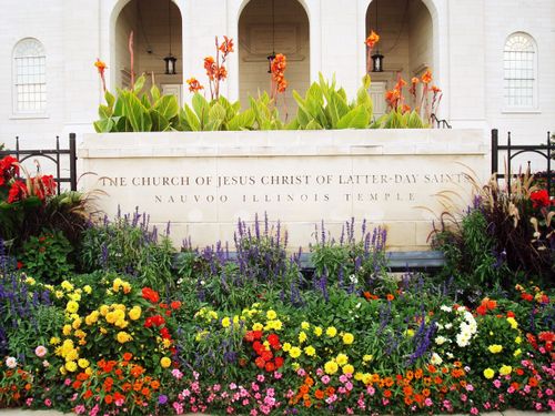 The stone sign outside of the Nauvoo Illinois Temple, surrounded by colorful flowers, with some of the temple’s arches seen in the distance.
