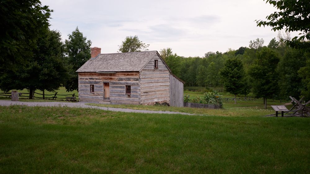 An exterior view of Joseph Smith's family exterior buildings in Palmyra, New York. 
There is farmland and a few smaller buildings on the land. They appear to be a barn and some kind of shed, likely another small barn or storage for a wagon.