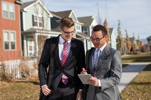 Two elder missionaries walk down a residential street while talking and looking at a small tablet.