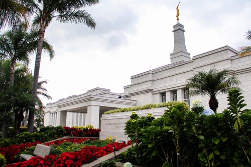 The entrance to the San José Costa Rica Temple, with a view of the temple name sign and the grounds around the temple. Trees, flowers, and bushes line the grounds.