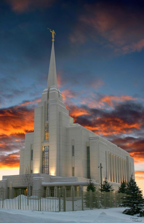 The entire Rexburg Idaho Temple lit up in the late evening, with snow on the grounds, a sunset in the background, and a fence around the temple grounds.