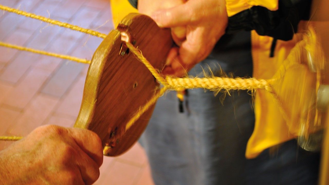 Two sets of hands holding a wooden rope making device, a yellow rope is wrapped around it.