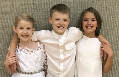 Three children stand together inside a meetinghouse.  The girls are in white...possibly at a baptism.The boy has his arms around the