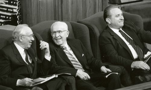 A black and white photo of Ezra Taft Benson, Gordon B. Hinckley and Thomas S. Monson smiling as they sit on the stand during General Conference.