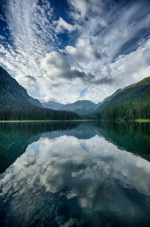 Clouds, mountains, and pine trees are reflected in a lake in Alaska.