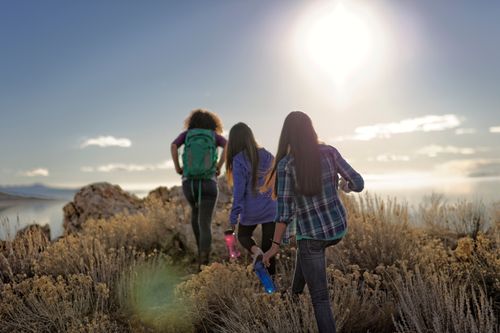 One young woman wearing a backpack and two other young women carrying water bottles hike together over a hill at girls’ camp.