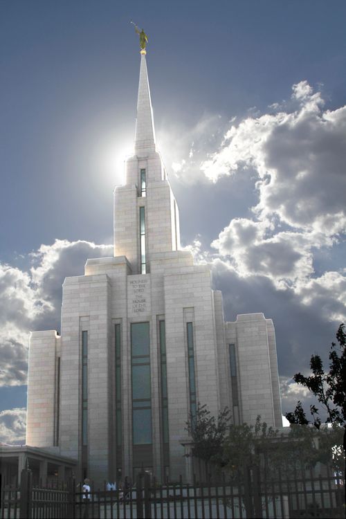 The front of the Oquirrh Mountain Utah Temple on a sunny day, with large white clouds and a ray of sun behind the temple’s spire.