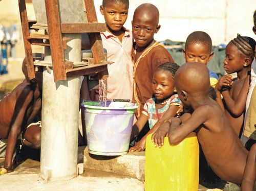 A group of African children standing closely by a tall water pump and watching clean water pour into a light purple bucket.