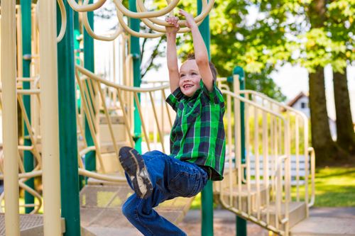 A little boy in a green and blue plaid shirt swings across monkey bars on a playground.