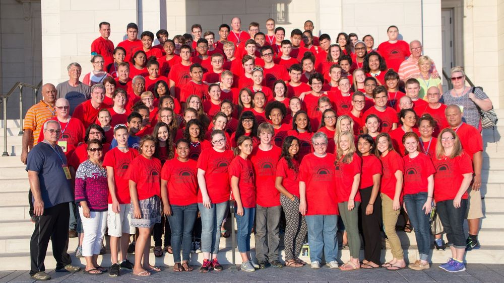 Over 100 youth and adult leaders, most in matching red T-shirts, pose for a large group photo on the front steps of a stone temple.