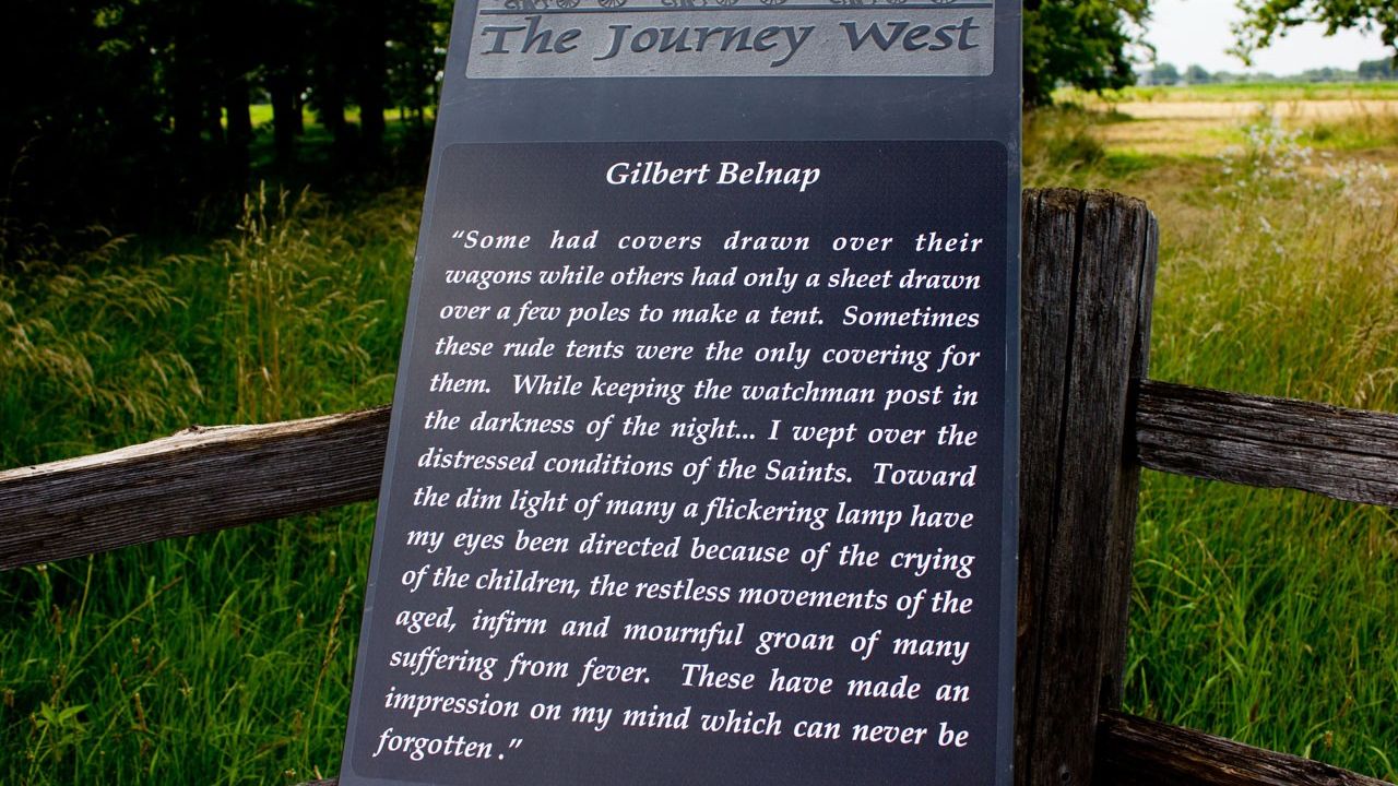 Close-up of interpretive marker with a wooden fence and grass field behind it. The sign is titled “The Journey West” and reads: “Gilbert Belnap ‘Some had covers drawn over their wagons while others only had a sheet drawn over a few poles to make a tent. Sometimes these rude tents were the only covering for them. While keeping the watchman post in the darkness of the night . . . I wept over the distressed conditions of the Saints. Toward the dim light of many a flickering lamp have my eyes been directed because of the crying of the children, the restless movements of the aged, infirm and mournful groan of many suffering from fever. These have made an impression on my mind which can never be forgotten.’”