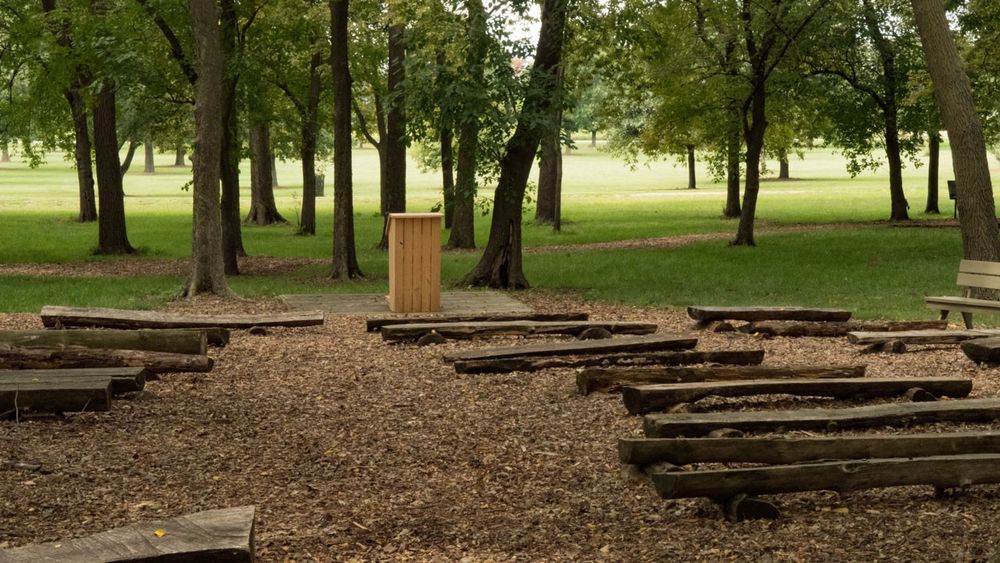 Log benches face a speaker’s podium in a grove of trees.