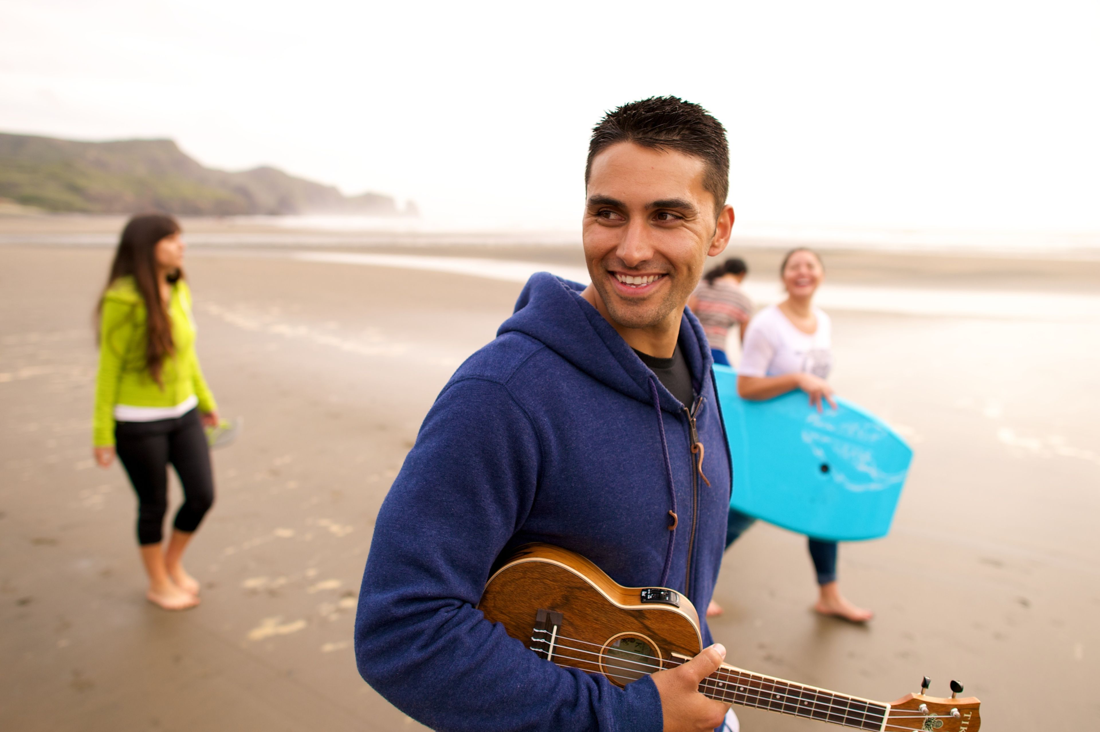 A young man walks along the beach, holding an ukulele, while other youth walk behind him.