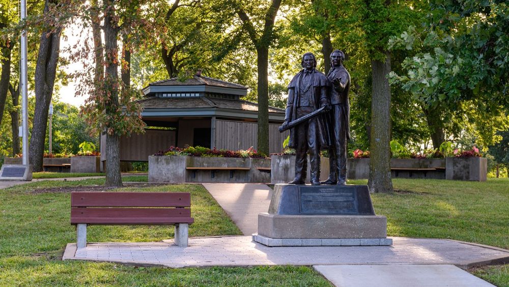 A bronze statue of two men with a wood pavilion and canopy of trees in the background.